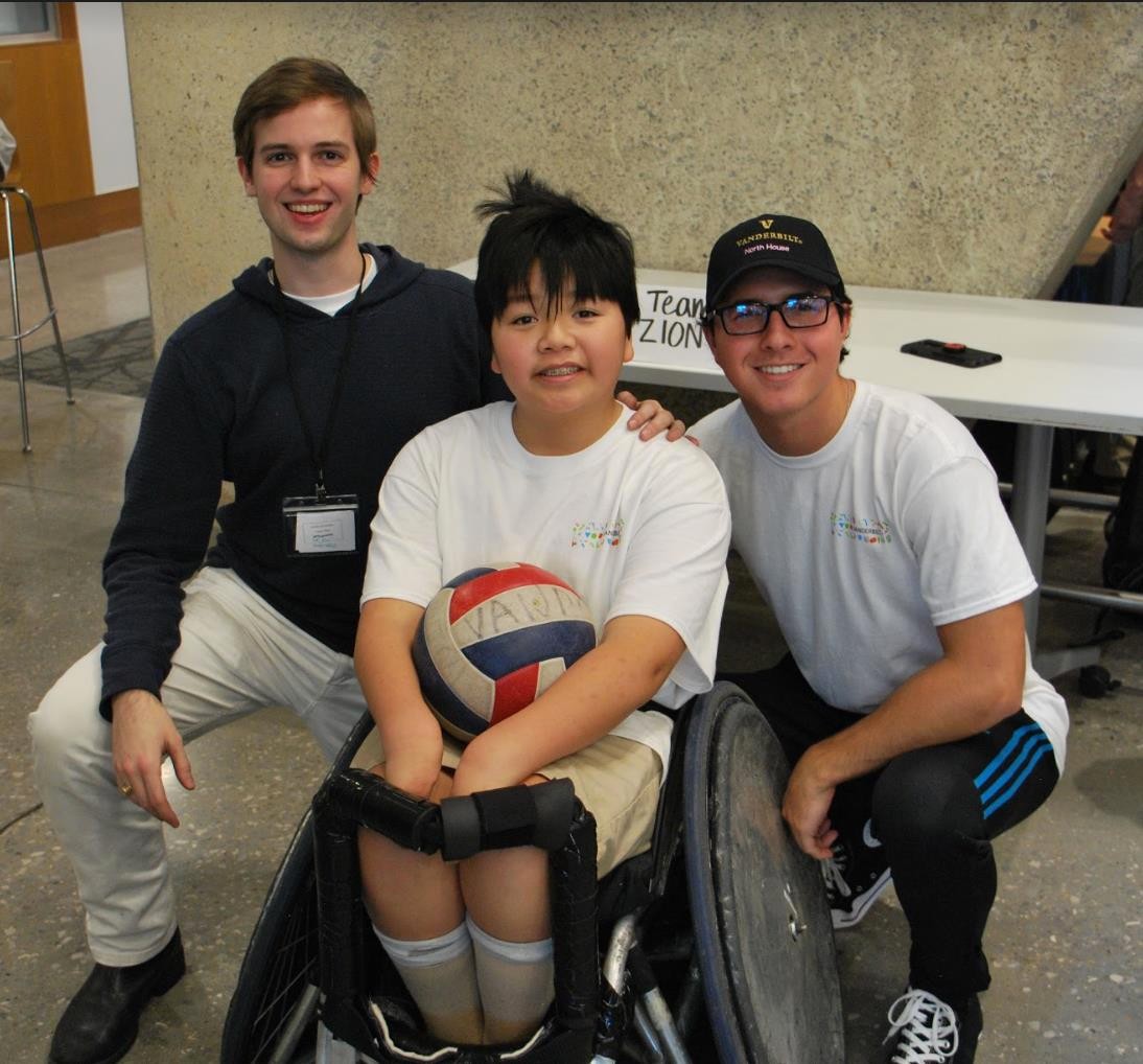 Tikkun Olam Team Zion Two college students are shown with a pre-teen or young teenager with a disability who is a wheelchair user