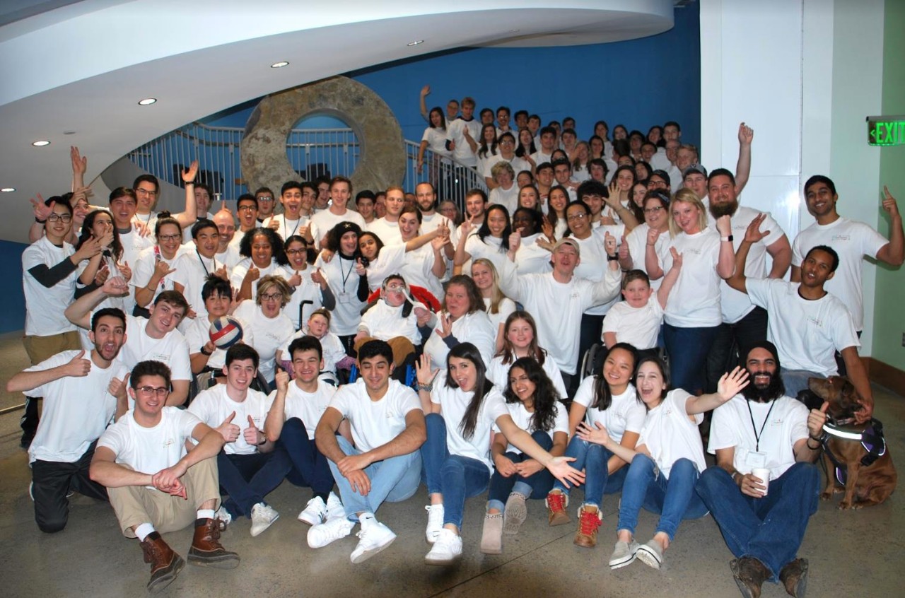 Tikkun Olam full group a large group photo of maybe 3 dozen people of all ages, all wearing matching white T-shirts, sitting and standing together, smiling and many are raising their hands in celebration. The group includes many college students, and children and adults with various types of disabilities and their families