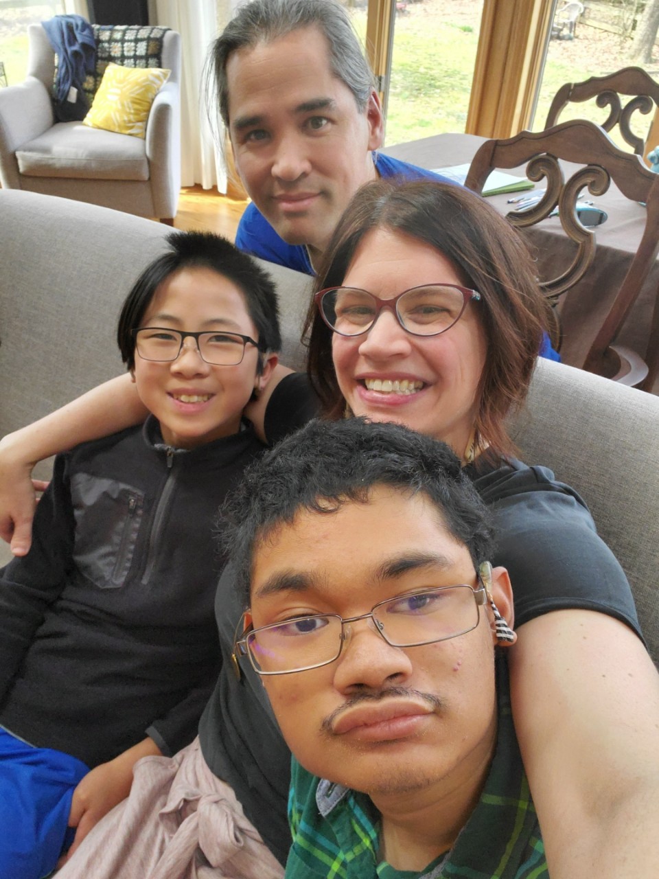 Guilaran family a family photo of the Guilarans – husband and father Fonsie is kneeling behind a couch where the rest of the family is sitting; the younger son Xiao Yu on one side of mom and wife Leslie, and the older teen son Angel on the other side; Leslie’s arms are around both of her sons. Leslie is a white woman with short brown hair, glasses and a big smile. Both sons are Southeast Asian and have short black hair and wire-rimmed glasses. Angel has a short mustache and a visible cochlear implant on one ear. Fonsie is Filipino and has long black hair pulled back in a ponytail