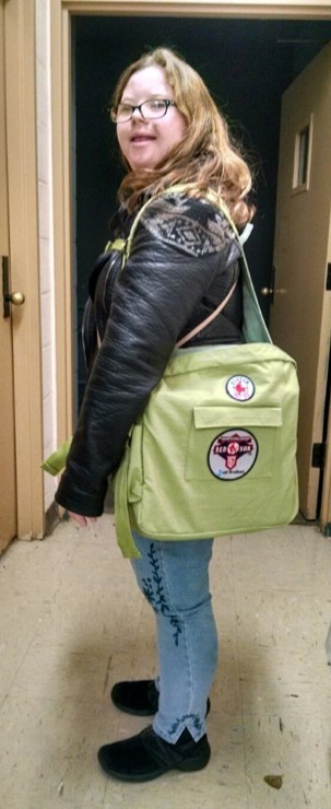A young white woman with Down syndrome, long brown curly hair, a leather jacker and glasses is shown in two poses, one from the side and one from the back, with her new custom backpack that distributes weight on the sides of her body instead of her back.