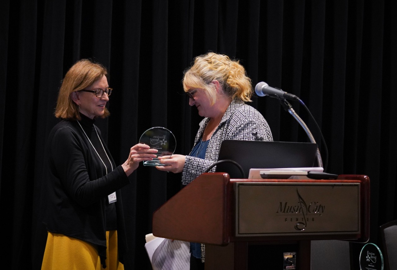 Wanda, wearing a black top and gold-colored skirt, accepts a round, glass award from Lisa Primm, a white woman with wavy blond hair and glasses, wearing a blue geometric blazer over a blue top. A large podium in the foreground, and the background is black curtains. 