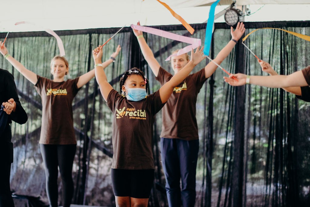 a young Black girl is on a stage with a few other dancers behind her, and all of the dancers are waving colorful ribbons on sticks in the air with their arms raised. They are all wearing matching shirts with the “MOVE” dance program logo that say “2021 Recital”.