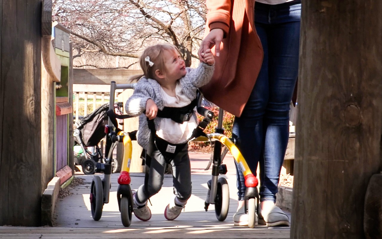 a very young little girl suspended by a walker, shown holding her mom’s hand as they walk together down the sidewalk. She is looking up at her mother and smiling.