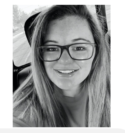 Black and white selfie photo of young white woman with black rimmed glasses and long straight hair, seated in a car