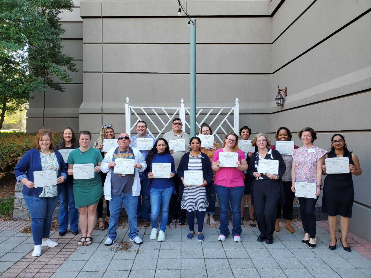 2023 Partners graduating class a diverse group of about 20 adults with and without disabilities pose for a group photo in the hotel courtyard holding up their Partners paper certificates.