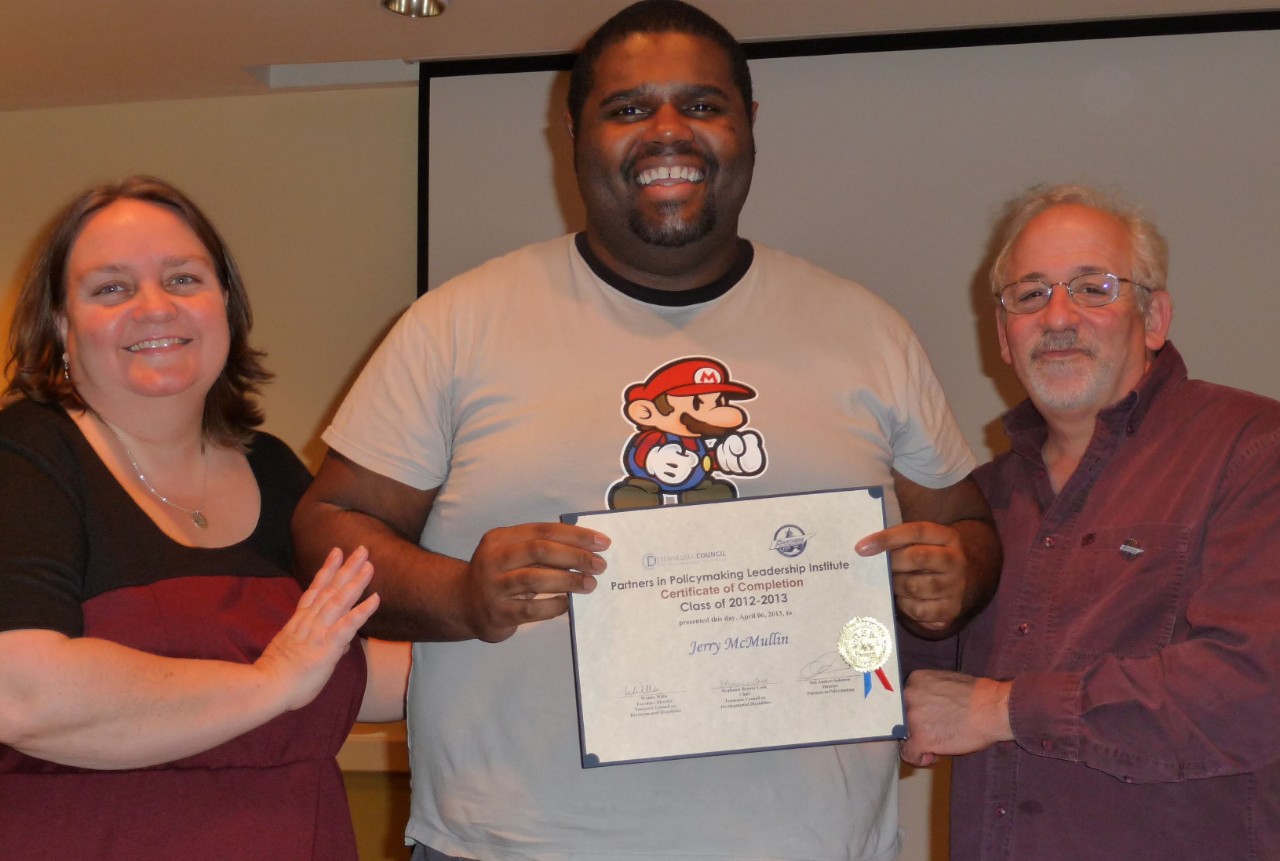Lynette Ned Jerry McMullin A white woman with shoulder-length brown hair, a Black man in a Mario shirt holding a certificate, and a white man with gray hair and beard and glasses smile at the camera.