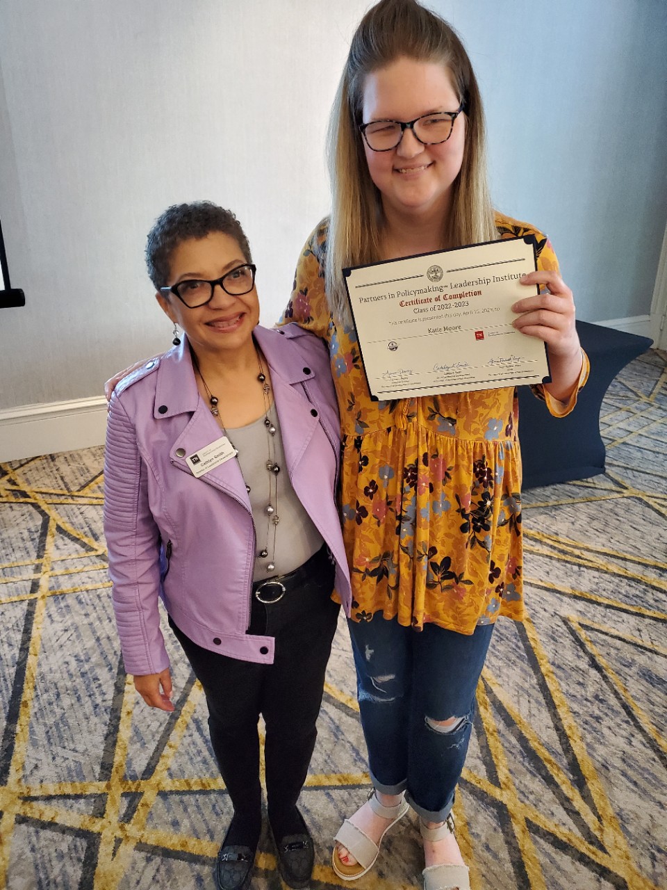 Cat and Katie Cathlyn Smith stands with her arm around Katie, a tall, blonde young woman in glasses and a yellow floral top and jeans, smiling and holding a certificate.