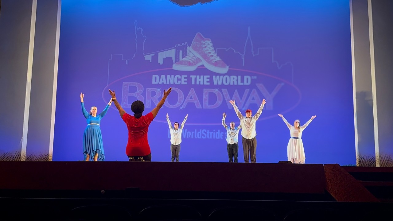 Borderless Arts TN dancers on a Broadway stage! Image description 1: Five adult dancers with disabilities stand on a Broadway stage with their hands raised in identical poses in the air; the 3 men are wearing slacks and button down white collared shirts; the two women are wearing similar flowing belted dresses, one blue and one white. Their instructor and choreographer has her back to the camera and is kneeling in front of the stage facing them. Behind the dancers, it shows a logo for “Dance the World: Broadway” which is a New York skyline and a pair of red sneakers.