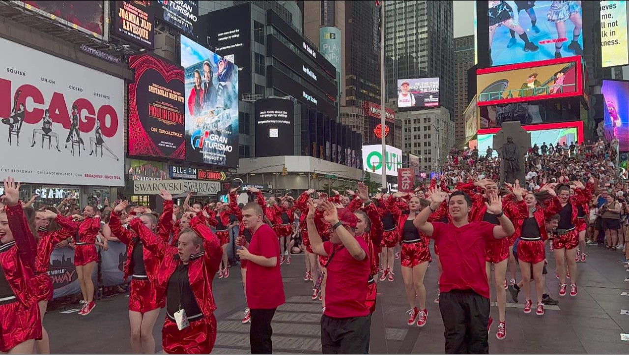Broadway 21 A large group of people are shown doing a choreographed dance down the middle of a large New York city street, all wearing bright red outfits.