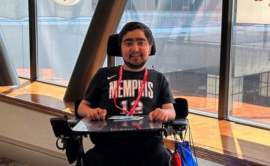 Image Description: Ravi Lipman, a person with medium-toned skin, sitting in a wheelchair and wearing a black t-shirt with the word "Memphis" on it. He is smiling and appears to be in an indoor setting with large windows behind him.