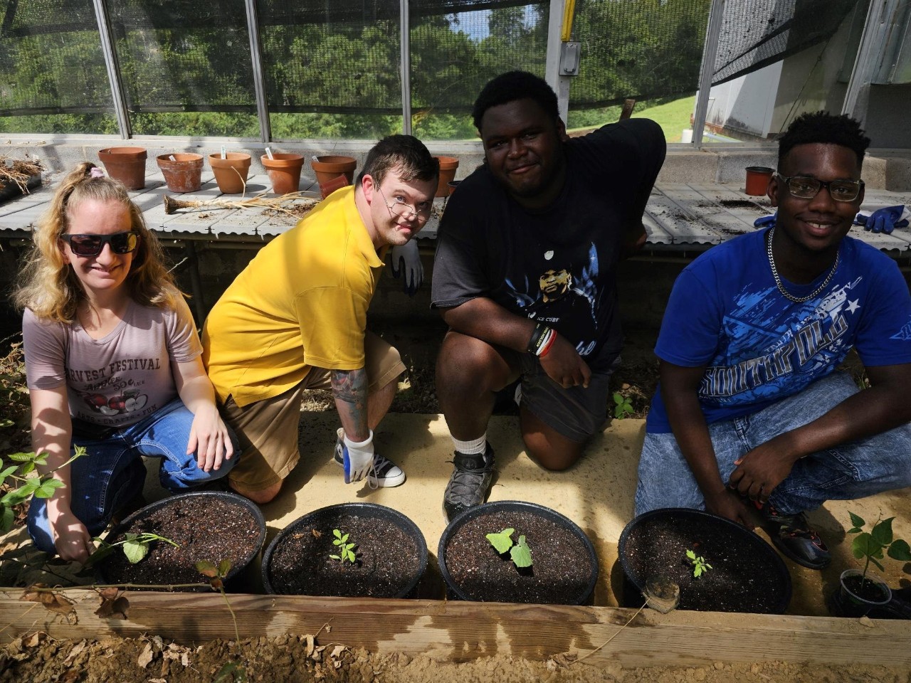 Image Description: Four people in a greenhouse setting, all smiling and engaged in plant care activities. From left to right: 1.	A light-skinned woman with blonde hair wearing sunglasses, a light-colored t-shirt with a graphic print, and blue jeans. 2.	A light-skinned man with short brown hair, glasses, and a yellow t-shirt, crouched down and smiling. 3.	A dark-skinned man with short black hair, wearing a black t-shirt with a graphic design and gray shorts, leaning forward with a relaxed smile. 4.	A dark-skinned man with short black hair, glasses, a blue t-shirt, and jeans, seated and smiling. Behind them are various potted plants and a greenhouse structure, providing a bright and green environment. They appear to be engaged in a hands-on learning activity related to plant care in a greenhouse.