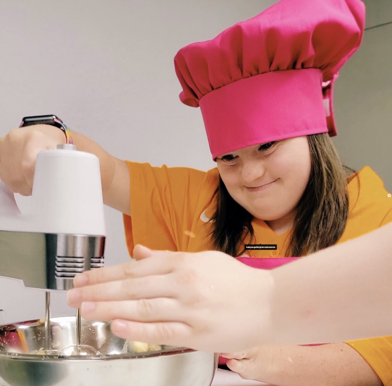 A light-skinned young girl wearing an orange shirt and a pink chef's hat uses an electric mixer to blend ingredients in a metal bowl. She is focused on her task, and her hands are steady on the mixer.