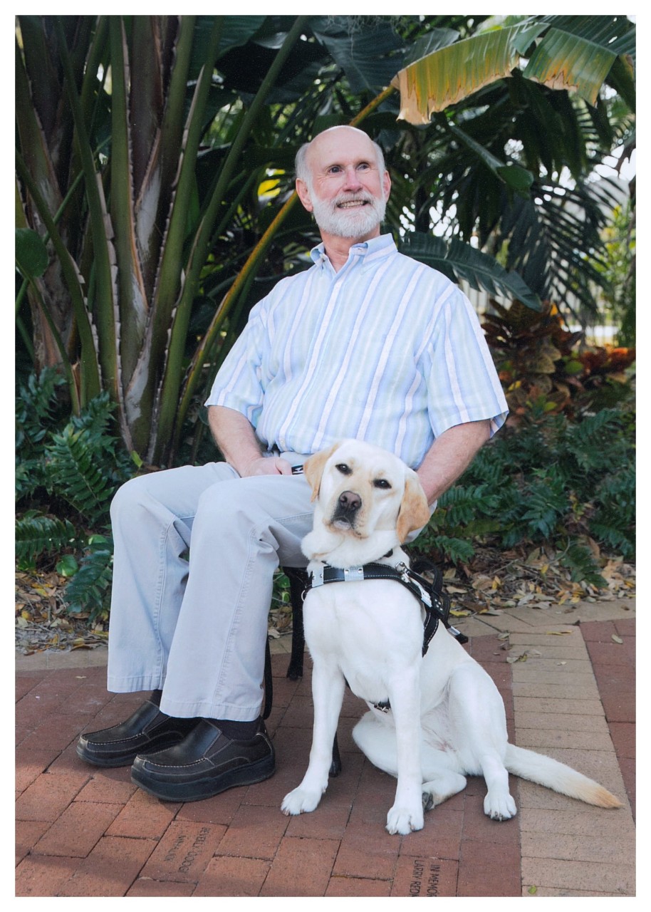 a gentleman with white hair and a beard is wearing a striped button up shirt and nice pants, sitting in a chair; next to him on the ground is a golden dog wearing a service dog vest