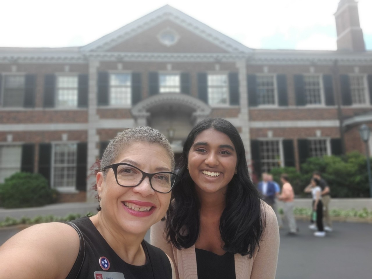 Selfie of two smiling women in front of a brick university building. Left: older woman with short curly hair, glasses, wearing black top with TN flag pin. Right: younger woman with long dark hair, wearing light cardigan. Background shows colonial-style architecture and people walking.