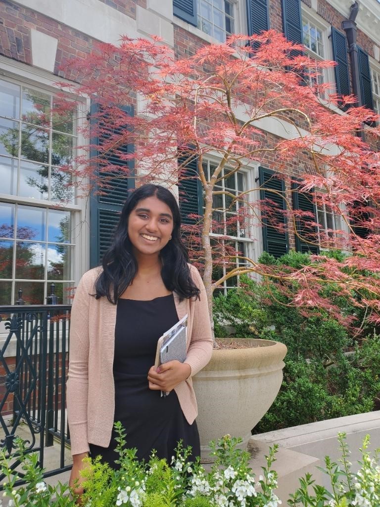 Eshani, who is a young woman, smiling at the camera. Eshani has long dark hair and brown skin. She is wearing a black top with a light pink cardigan and is holding what appears to be a notebook or folder. Eshani is standing in front of a brick building with large windows and black shutters, with a vibrant red Japanese maple tree in full foliage behind her.