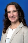 headshot of Jolene Sharp, a white woman with long curly brown hair, a white blouse and a beige suit jacket