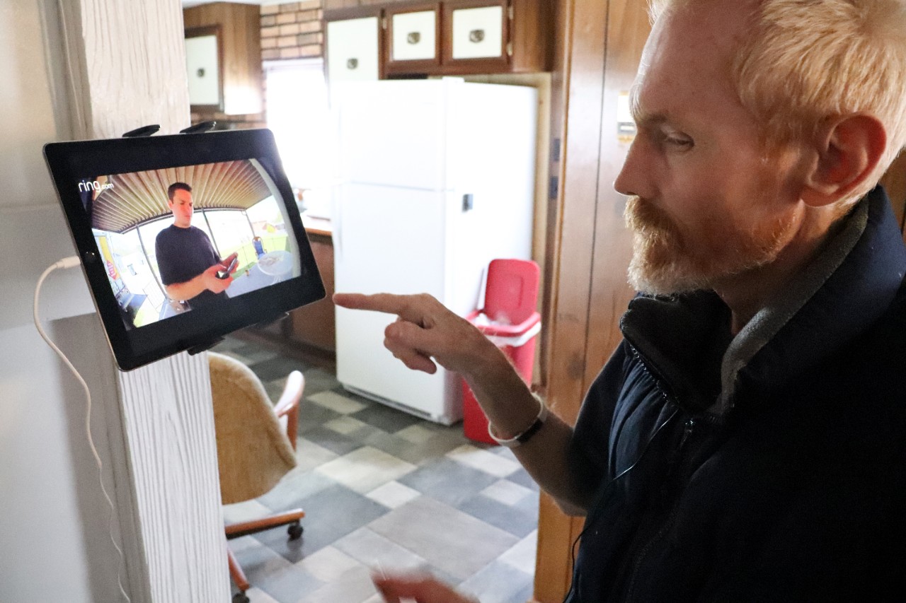 Carl using Enabling Technology an older white man with a red-blonde hair and beard and sweater in his home kitchen, looking at a tablet mounted on his wall that is showing him who is at his front door (it says the brand name “ring” in the corner of the video on the tablet). His name is Carl and he is about to click the tablet to allow his visitor into his home, after he saw who is at his door