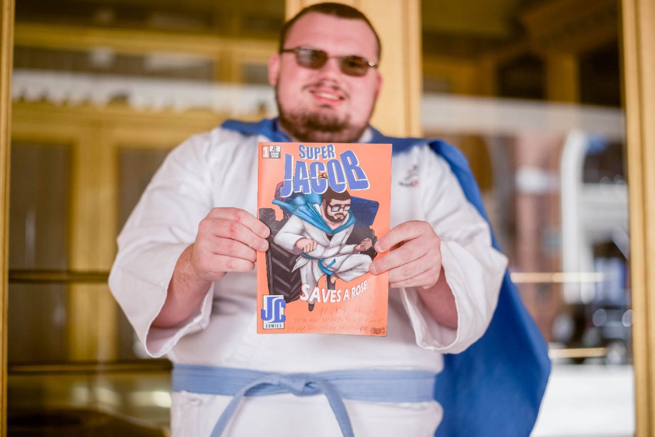 Jacob is holding out his comic book Super Jacob. The comic book has an image of a superhero wearing a white costume with a blue belt and cape
