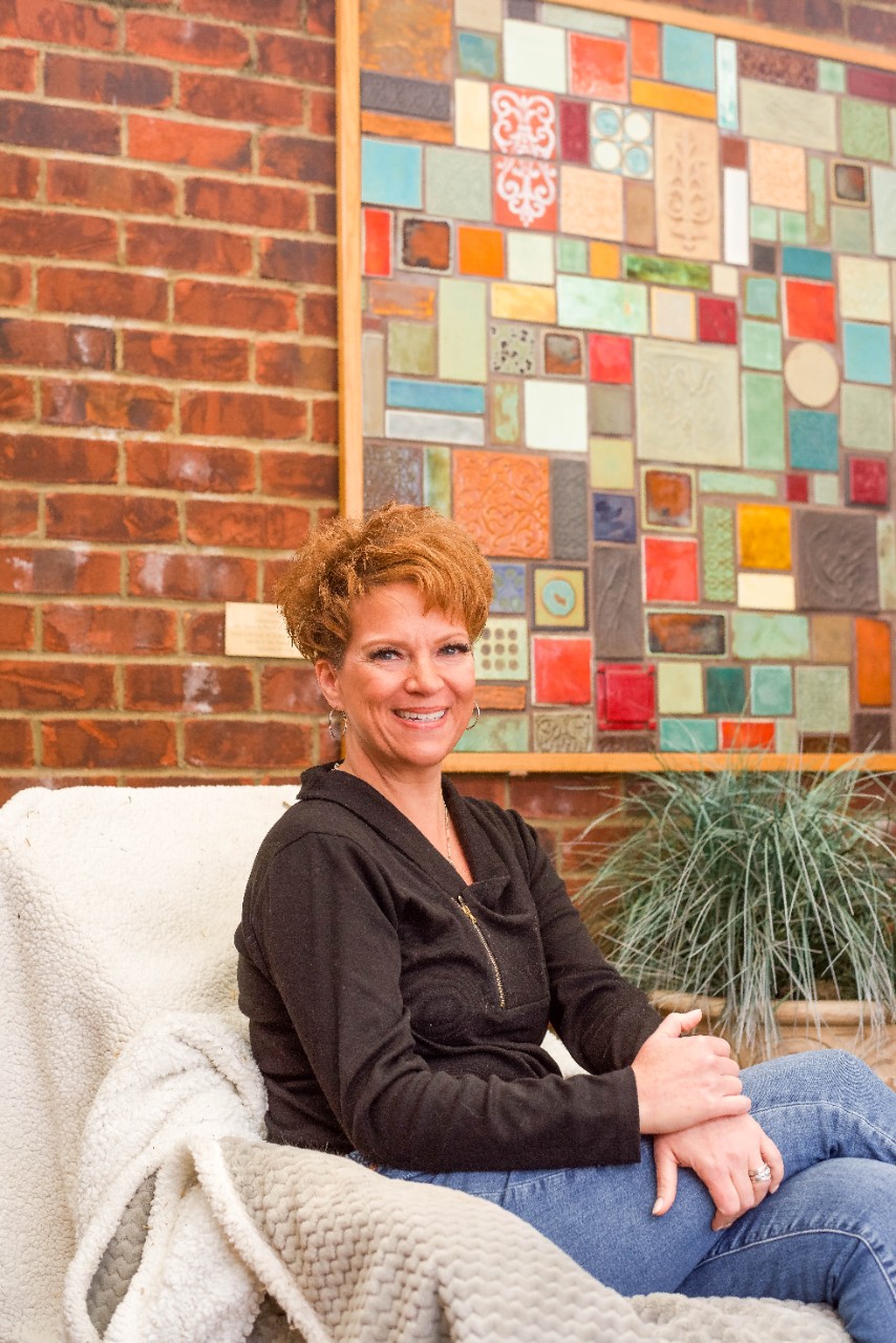 Juli, a 51 yo woman with short red hair, sits in front of a colorful mosaic painting outdoors