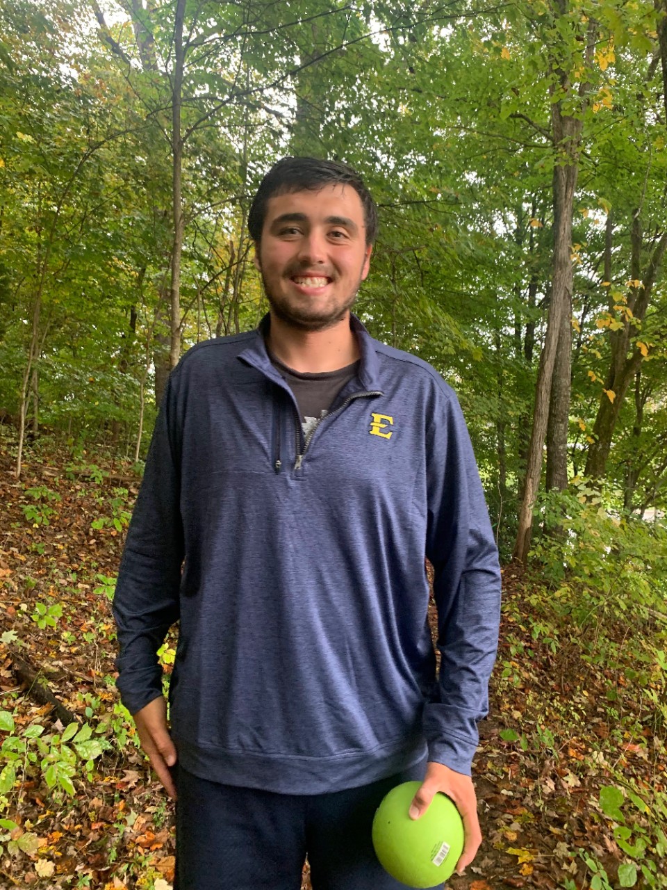 A medium complected man in an ETSU polo smiling outside and holding a green ball